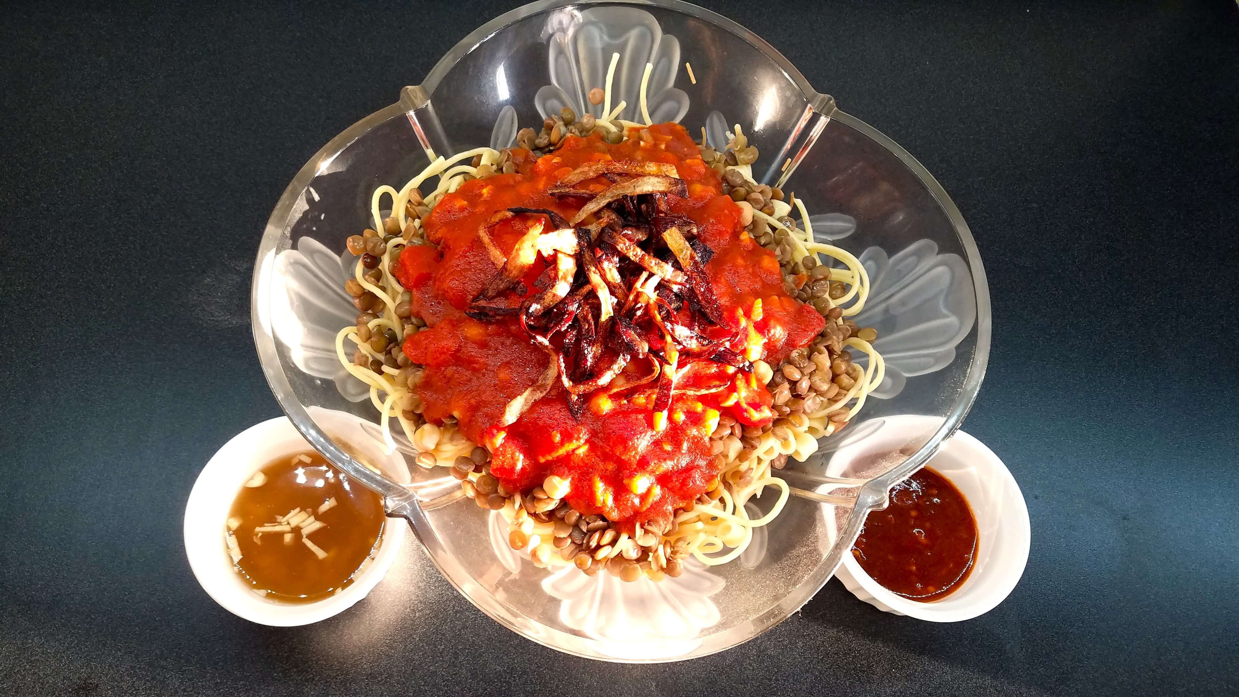Homemade Egyptian Koshari bowl featuring layers of toasted vermicelli rice, macaroni, brown lentils, and savory spiced garlic tomato sauce under crispy golden fried onion.