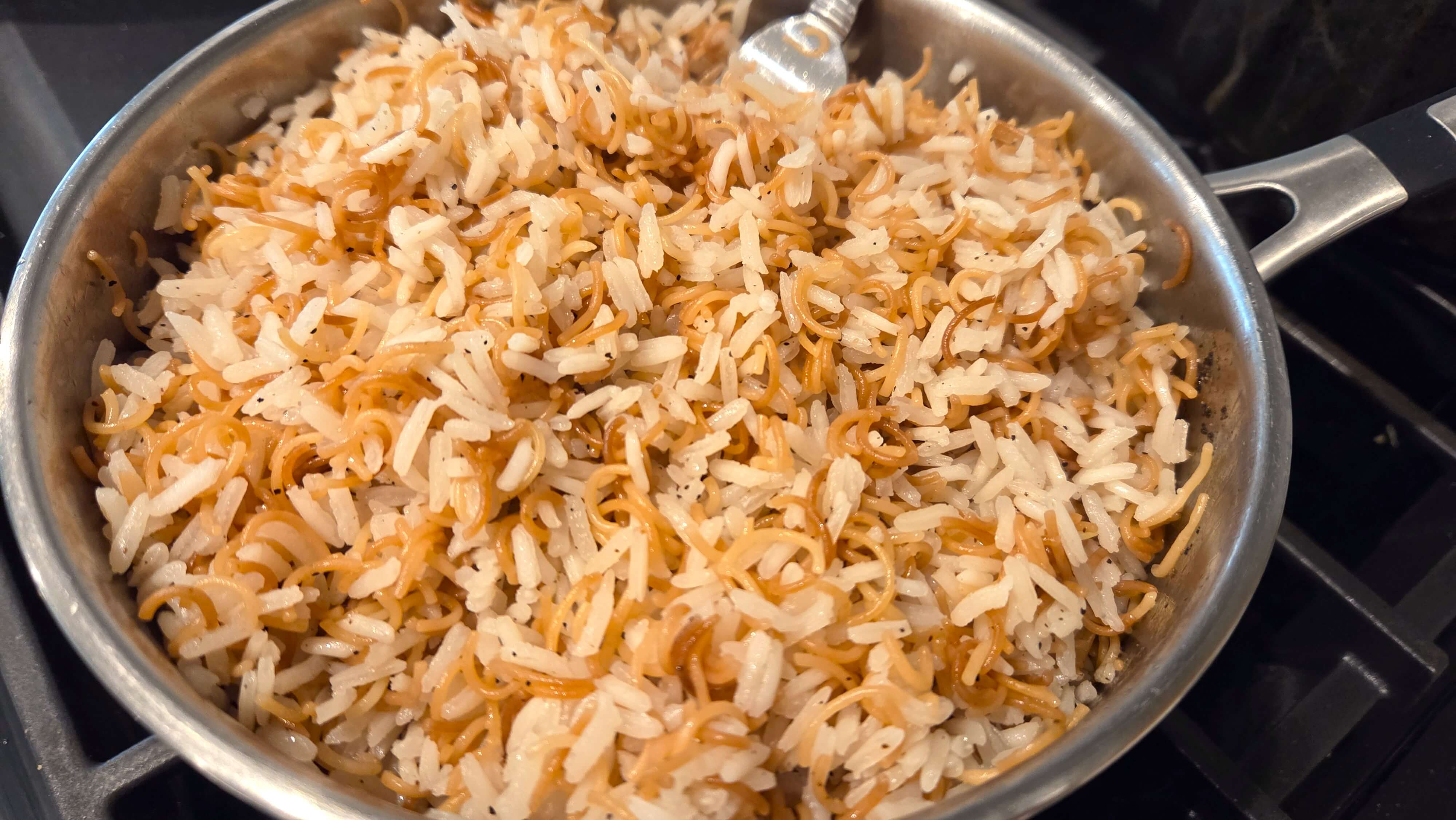 Gently fluffing the fully cooked, non-sticky Egyptian vermicelli rice with a metal fork to separate the grains.