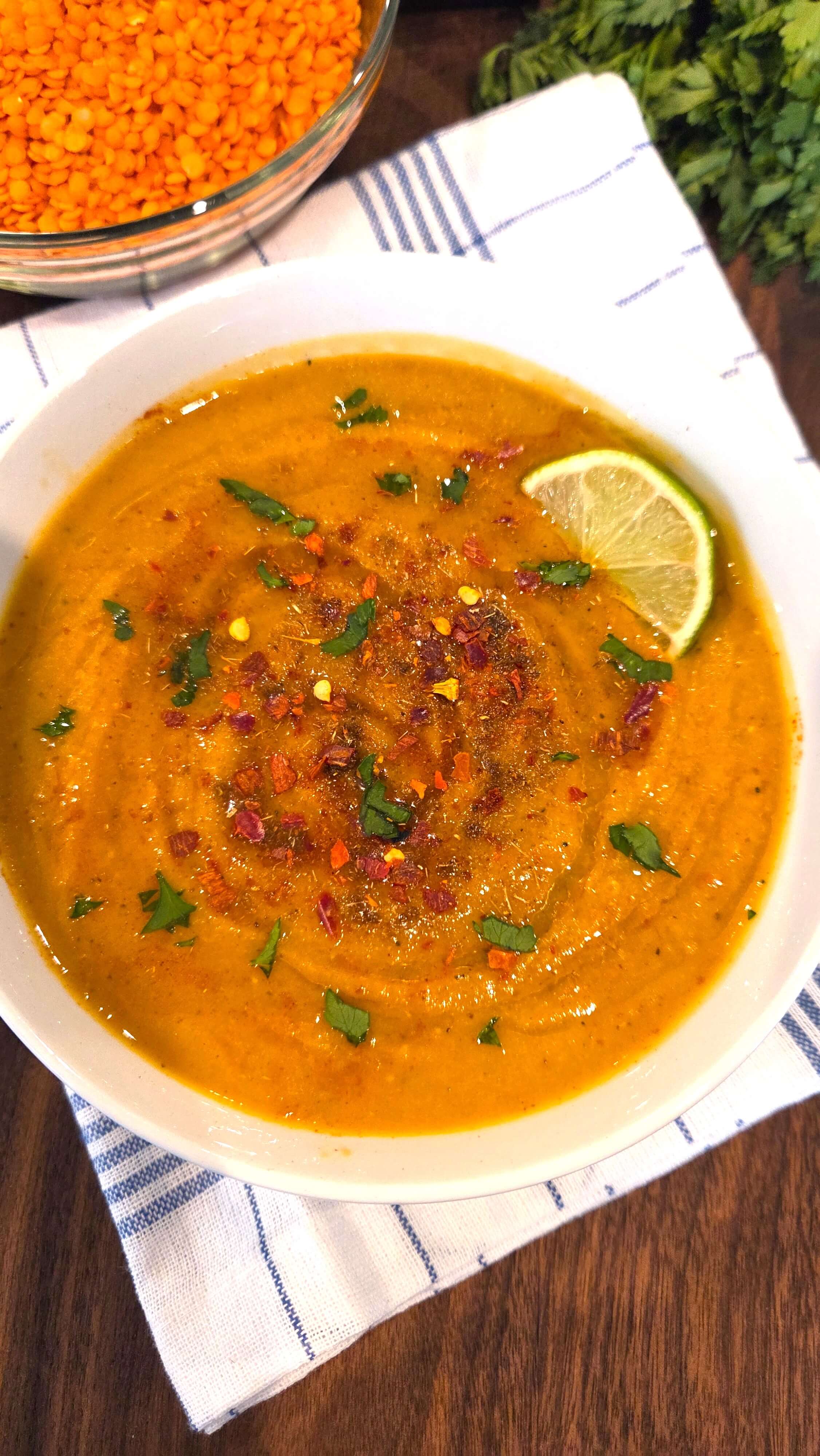 A top-view angle shot of a cozy bowl of authentic Egyptian lentil soup (Shorbat Ads) with fresh parsley, paprika, cumin, red pepper flakes, and lemon wedge.