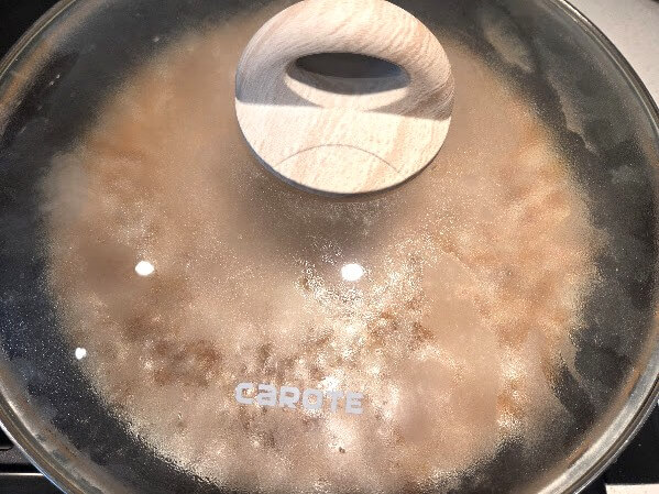 A pot sitting on a stove burner with a glass lid tightly sealed to trap the steam while the rice simmers.