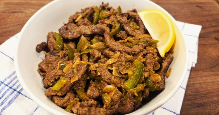 A wide overhead shot of a sizzling iron skillet filled with hot Egyptian Kebda Eskandarani beef liver strips and sliced green chili peppers.