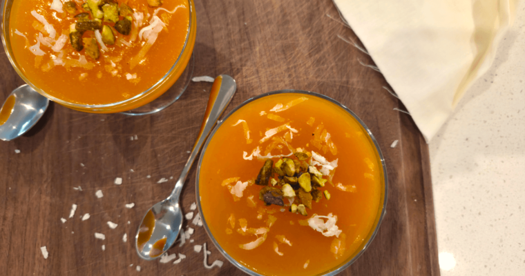 A wide overhead shot of several chilled glass dessert bowls filled with vibrant orange Qamar Al Din pudding garnished with shredded coconut and crushed pistachios.