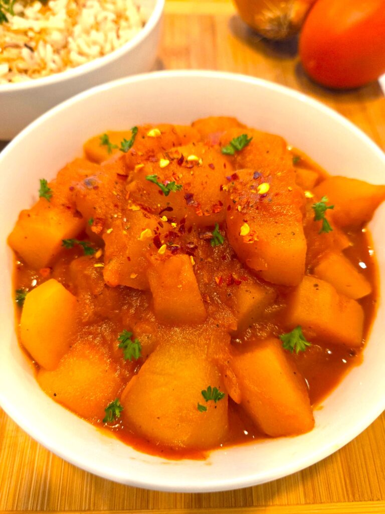 A beautiful close-up of a single comforting bowl of Egyptian potato stew served hot next to a platter of traditional rice with vermicelli, highlighting the rich tomato sauce coating.