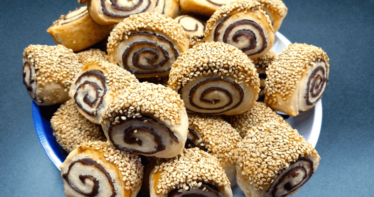 A wide overhead shot of a festive holiday platter filled with golden brown vegan date pinwheel cookies, displaying clean dark date spirals and crunchy raw sesame seed borders.