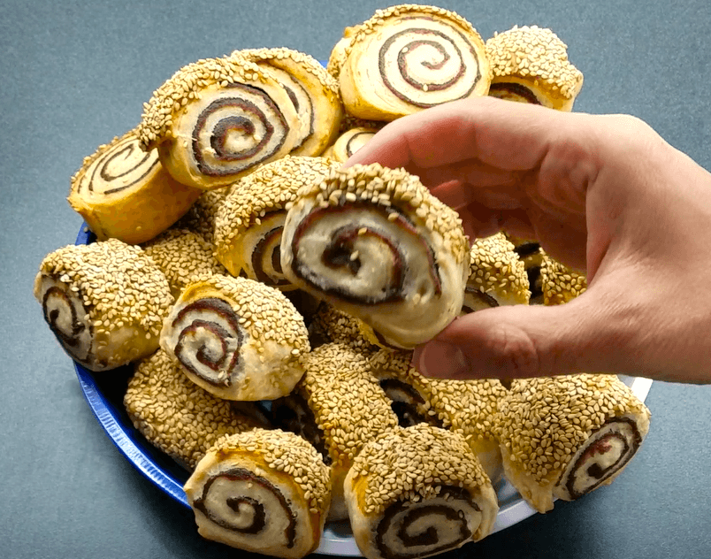 A premium close-up shot of a single soft vegan date pinwheel cookie, showcasing the perfect contrast of the dark sweet date paste spiral and the toasted crunchy sesame seed crust.