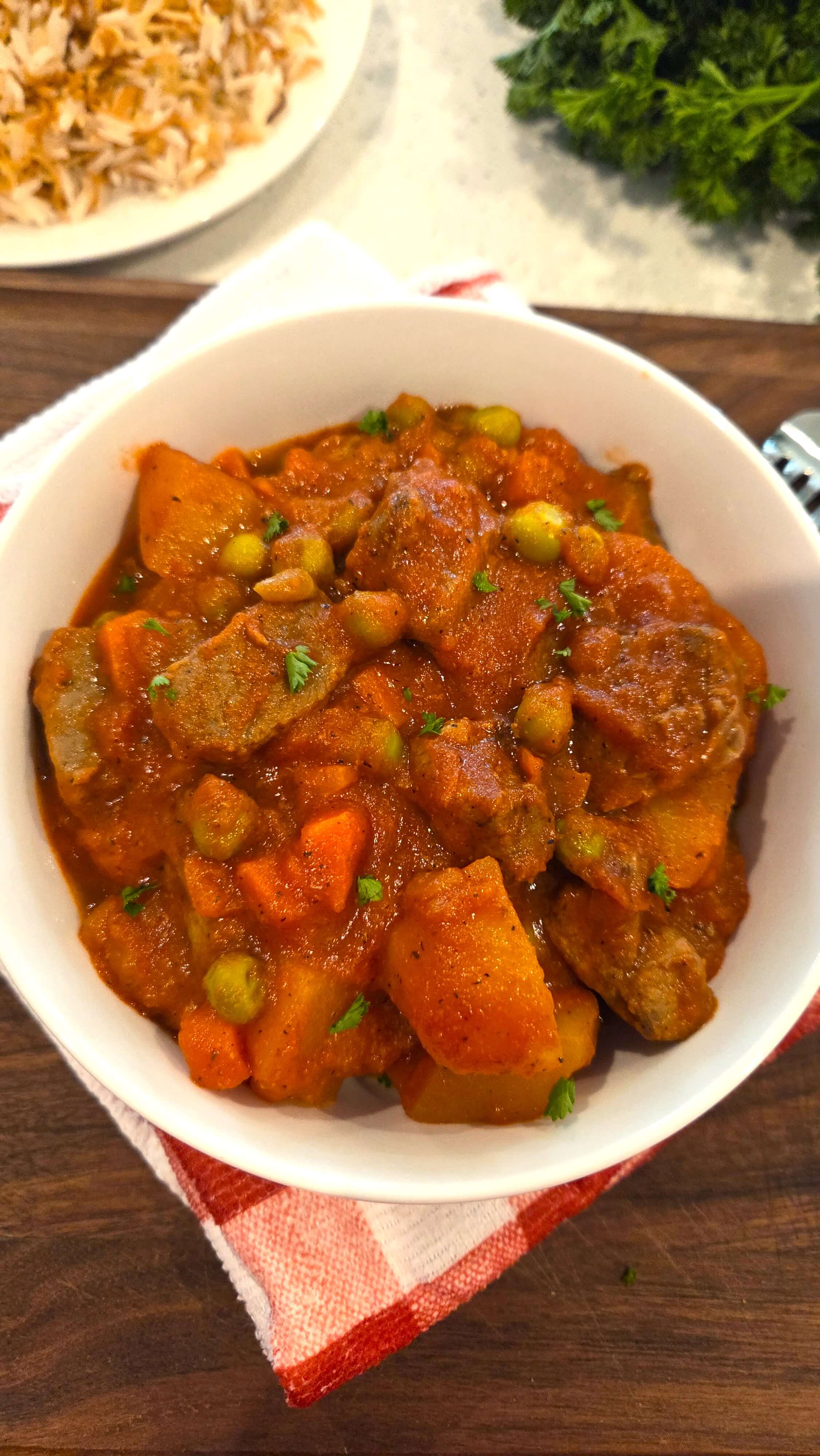 A premium close-up shot of a steaming bowl of homemade Egyptian Bazella stew, showing tender beef cubes, peas, carrots, and potatoes in a velvety tomato gravy.