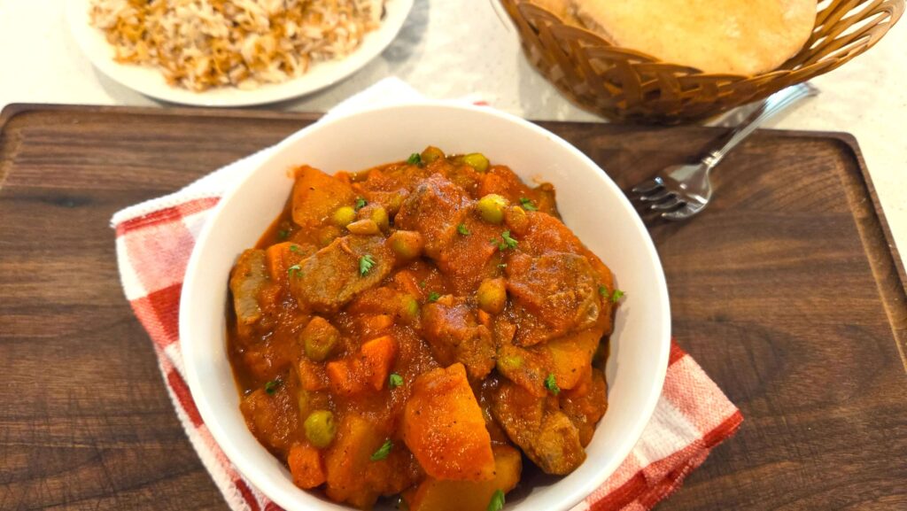 A beautiful top-down shot of a bowl of thick peas and carrots stew with beef chunks, served hot alongside a platter of Egyptian vermicelli rice and pita bread.