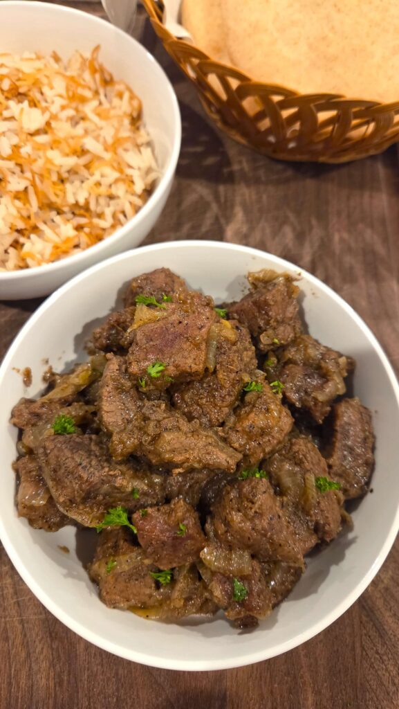 A beautiful close-up of a single comforting bowl of Egyptian Kabab Halla beef served hot next to a platter of traditional rice with vermicelli and warm pita bread.