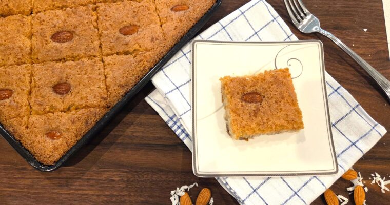 A wide overhead shot of of a sliced diamond piece of Egyptian vegan basbousa in a white ceramic plate next to a big tray of basbousa cut into diamond shapes.