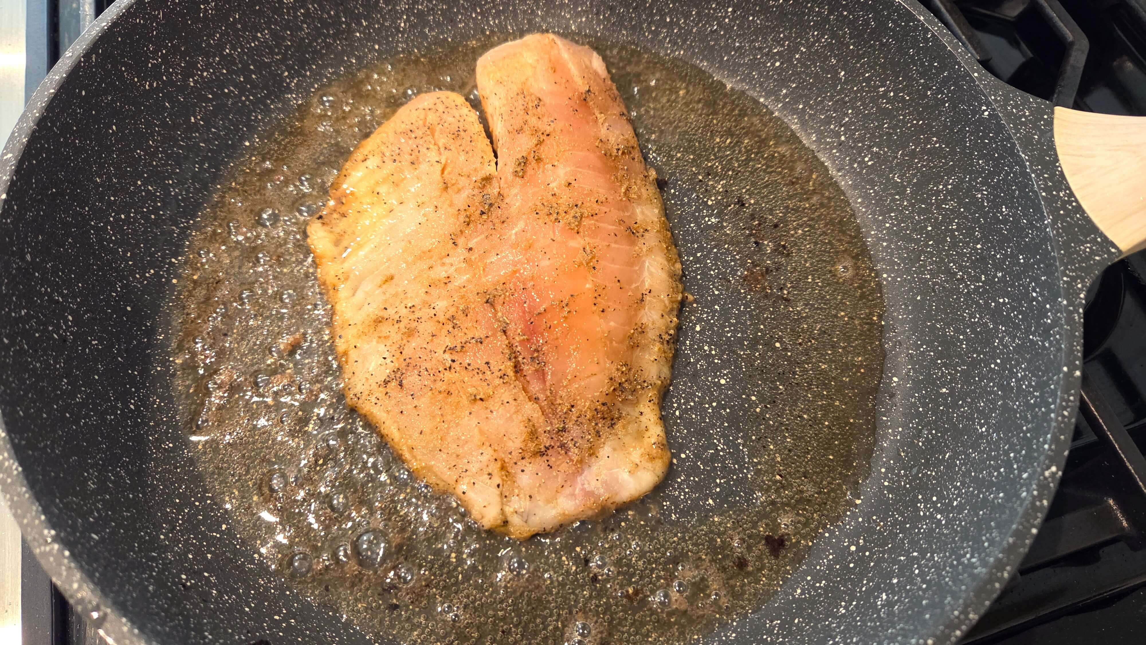 Tilapia fillets cooking in olive oil in a skillet until golden on the first side.
